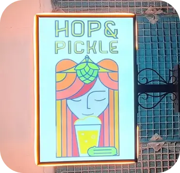 Illuminated rectangular blade sign for Hop & Pickle displaying custom artistic graphics, representing creative commercial building signage.